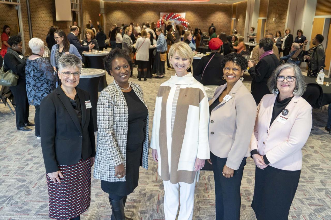 Five women senior leaders standing in a group with people mingling behind them.