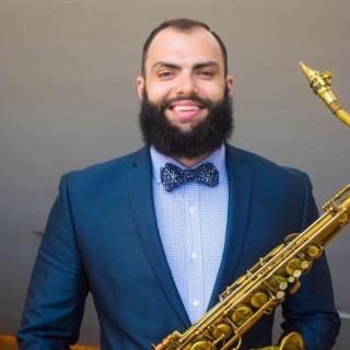 Robert Brooks wearing a blue suit with a bowtie and holding a saxophone against a grey background. 