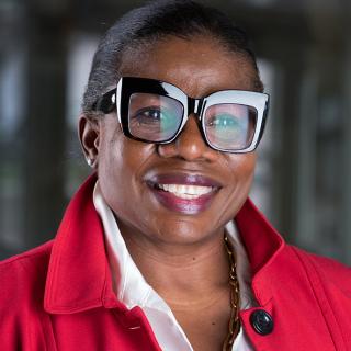 Portrait of Ruby Pentsil-Bukari, Black woman wearing a red jacket, gold chain and large glasses